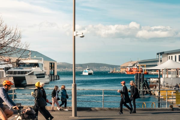 A photograph of mixed modes of transport along Hobart's waterfont, facing out towards the Derwent, including people walking in both directions, a cyclist, and a ferry about to dock.